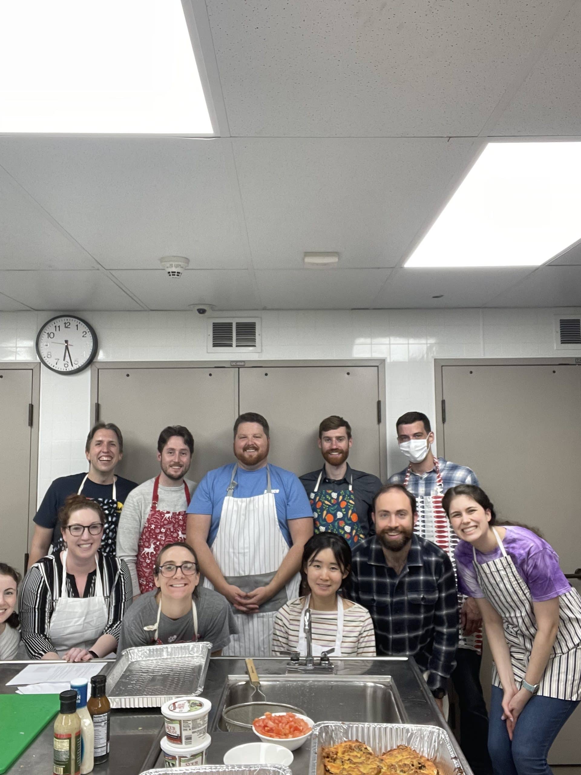 A group of people in their 20s & 30s,most wearing aprons over their clothes, smile together in a kitchen.