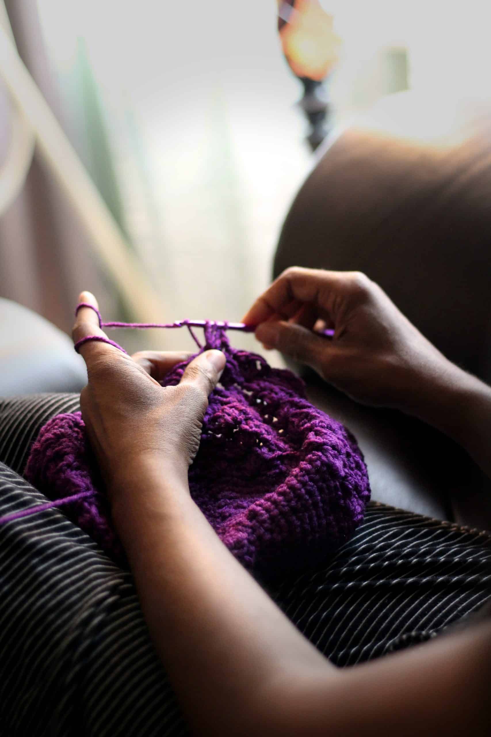 A Black woman's hands work with purple yarn onto a needle. She is creating some kind of fiber garment, scarf, or hat.