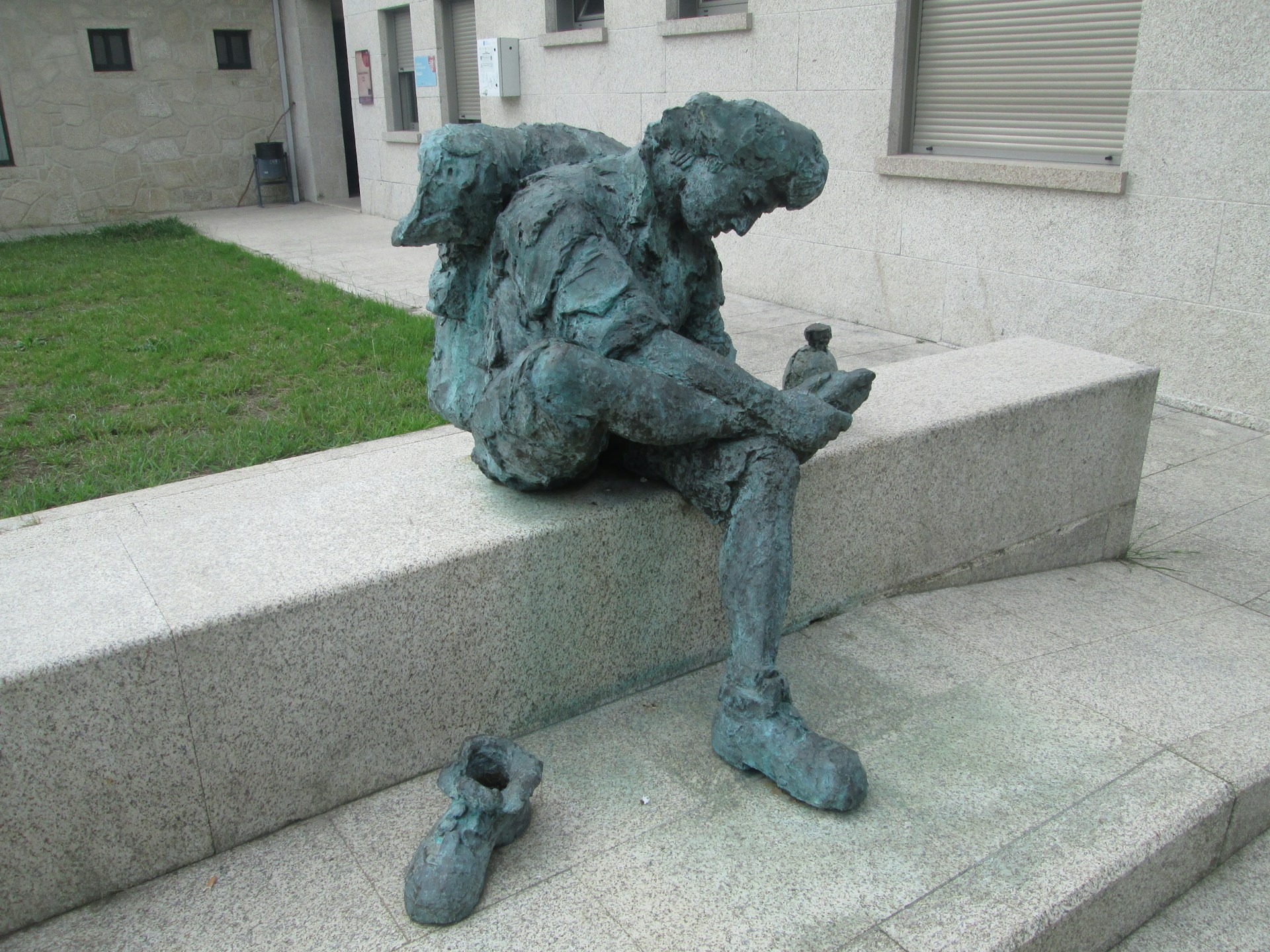 a greenish patina-covered sculpture depicts a person sitting on a wall and checking the underside of their foot, with a separate small show scuplture close by. It is associated with the Camino Santiago pilgrimage in Spain.